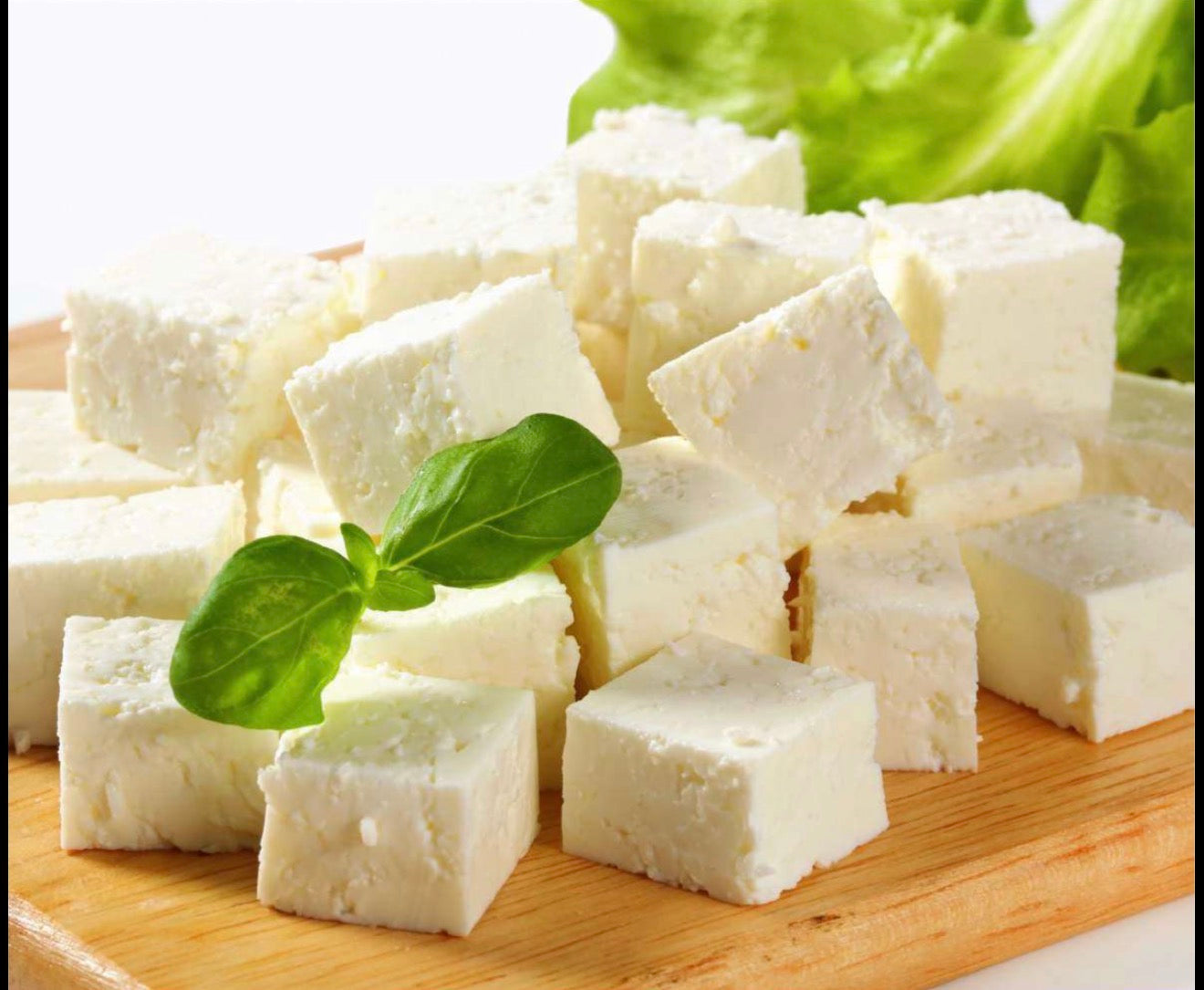 Cubes of fresh feta cheese arranged on a wooden cutting board with basil leaves and lettuce in the background.