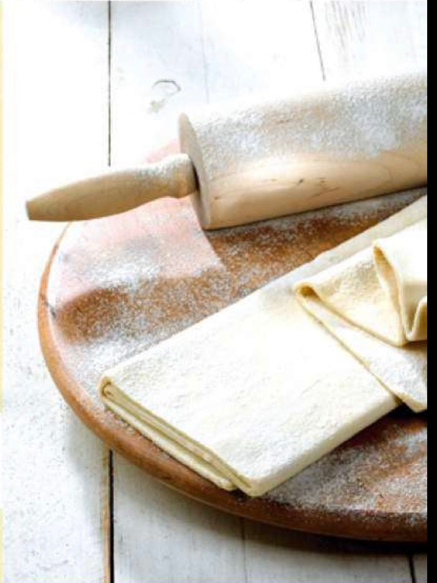 Wooden rolling pin and folded sheets of puff pastry dusted with flour on a round wooden board.