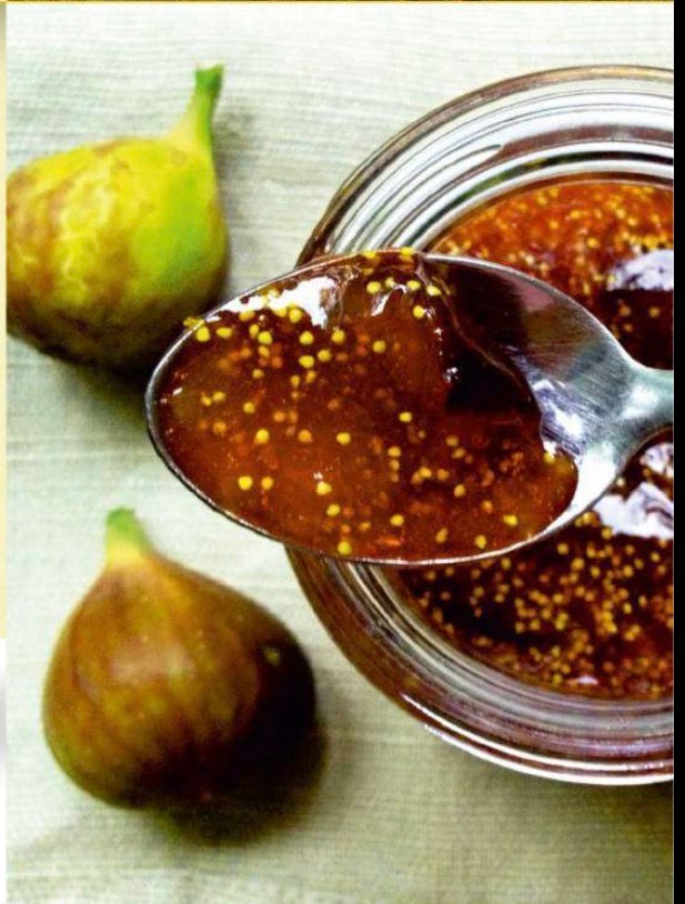 Spoonful of fig jam above a glass jar with whole fresh figs beside it on a light fabric surface.