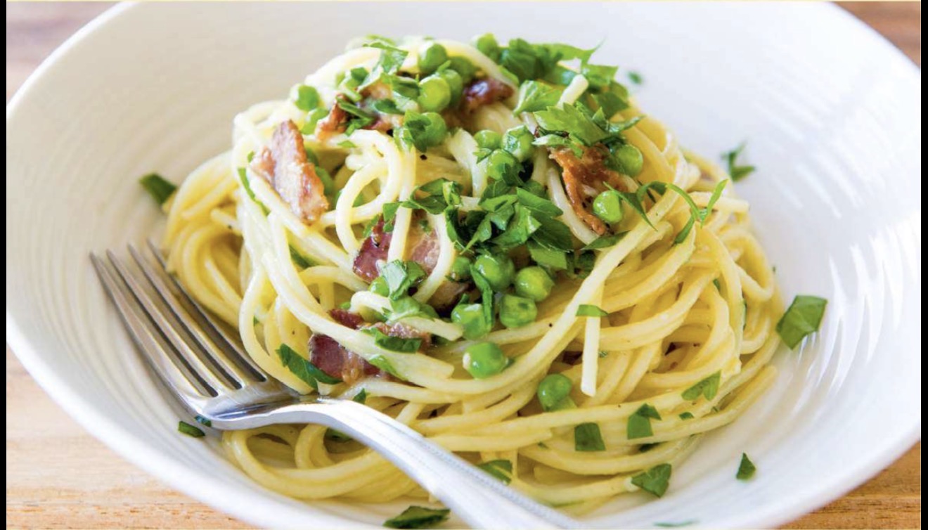 Bowl of spaghetti pasta topped with green peas, crispy bacon pieces, and fresh chopped herbs with a fork on the side.