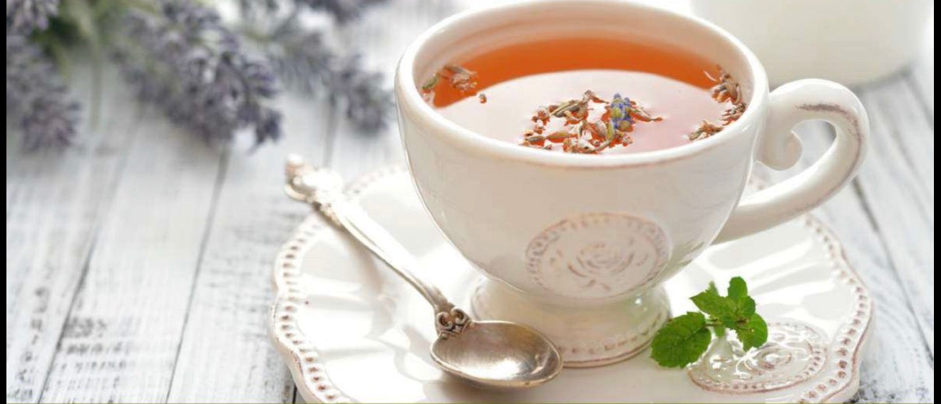 White teacup filled with herbal tea and flower petals on a matching saucer, with a silver spoon and fresh mint leaves beside it.