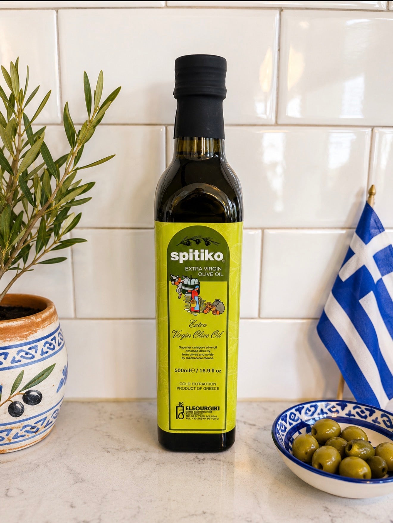 Bottle of Spitiko olive oil on a kitchen counter with a Greek flag and olive branch in the background.