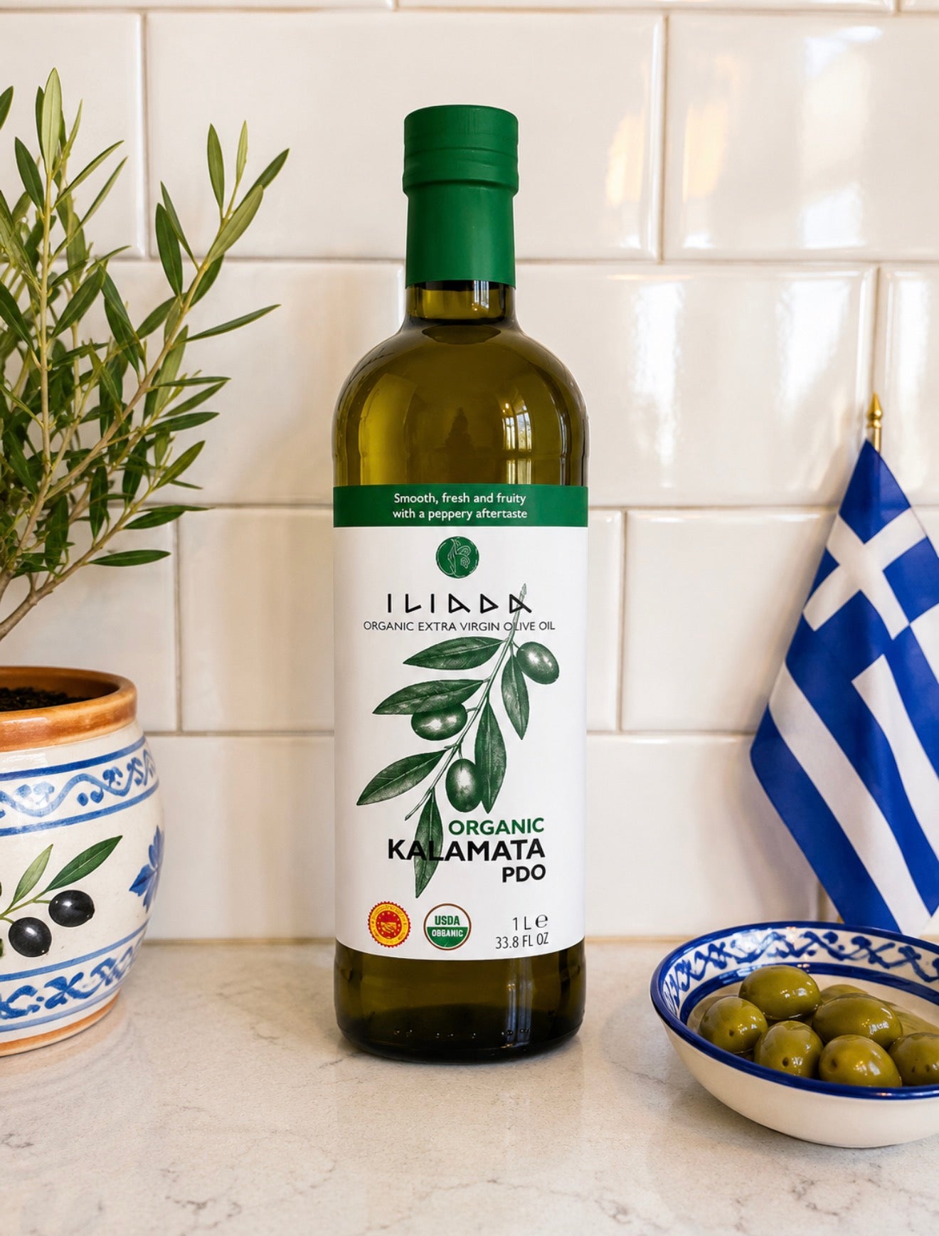 Bottle of Iliada Organic Kalamata PDO olive oil on a kitchen counter with a Greek flag and bowl of olives.